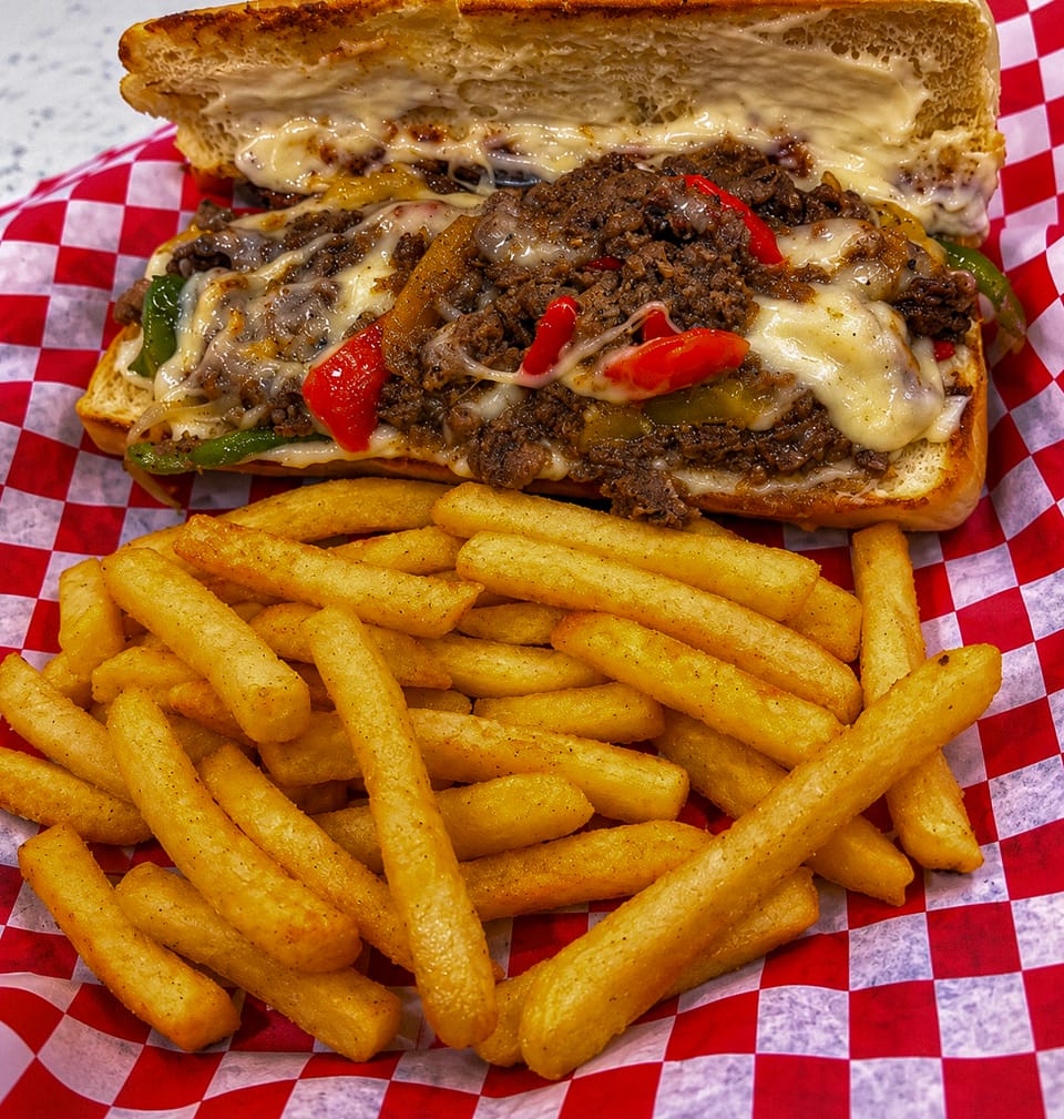 A toasted roll piled with seasoned beef, peppers and melted cheese, with fries, on red-and-white checkered paper